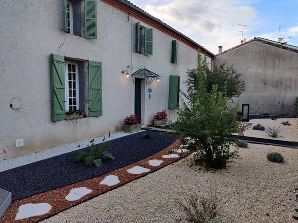 A rustic building with green shutters, a gravel pathway, and potted plants.