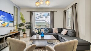 Ein modernes Wohnzimmer mit Sofa, Couchtisch und Sesseln. Links befindet sich ein Fernseher und ein großes Fenster mit Vorhängen.