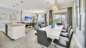 Eine moderne Küche mit einer zentralen Kochinsel, einem Essbereich mit langem Tisch und Stühlen sowie einem großen Fenster mit Vorhängen.