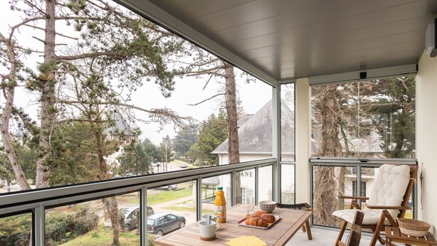 A balcony with a wooden table set for breakfast, a cushioned chair, and a view of trees and a car in the background.