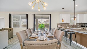 A modern kitchen with a dining area, featuring a large island, pendant lighting, and a view of the outdoors.