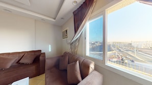 A living room with a large window, a brown sofa, and a view of a cityscape. - Hotel Al Madinah Hotel Apartments (Madinah)