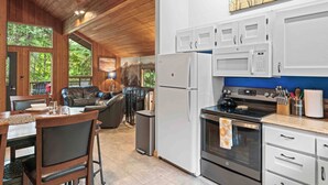 Una cocina moderna con armarios blancos, horno de acero inoxidable y refrigerador. Hay un comedor con mesa y sillas, y una sala de estar con sofá y chimenea.