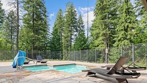Una piscina con sillones de descanso y una zona cubierta, rodeada por una valla y árboles altos.