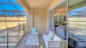 Un balcón con un sofá blanco, una mesita y una silla. Hay una puerta y un muro de cristal con vistas a una piscina y edificios.