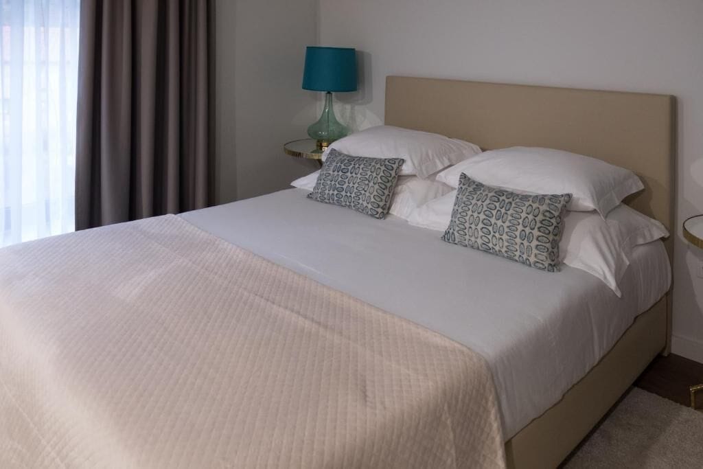 Una cama bien hecha con una colcha blanca y dos almohadas estampadas, una lámpara turquesa sobre una mesita de noche y vistas a una ventana con cortinas.