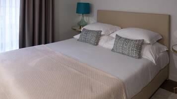 Una cama bien hecha con una colcha blanca y dos almohadas estampadas, una lámpara turquesa sobre una mesita de noche y vistas a una ventana con cortinas.