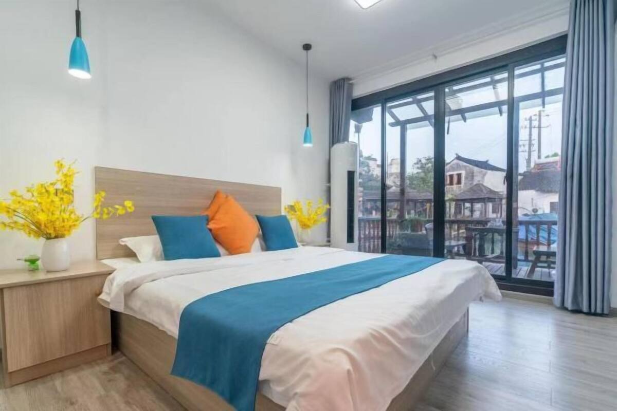 A modern hotel room with a large bed, a nightstand, a vase with yellow flowers, and a view of buildings through large windows.