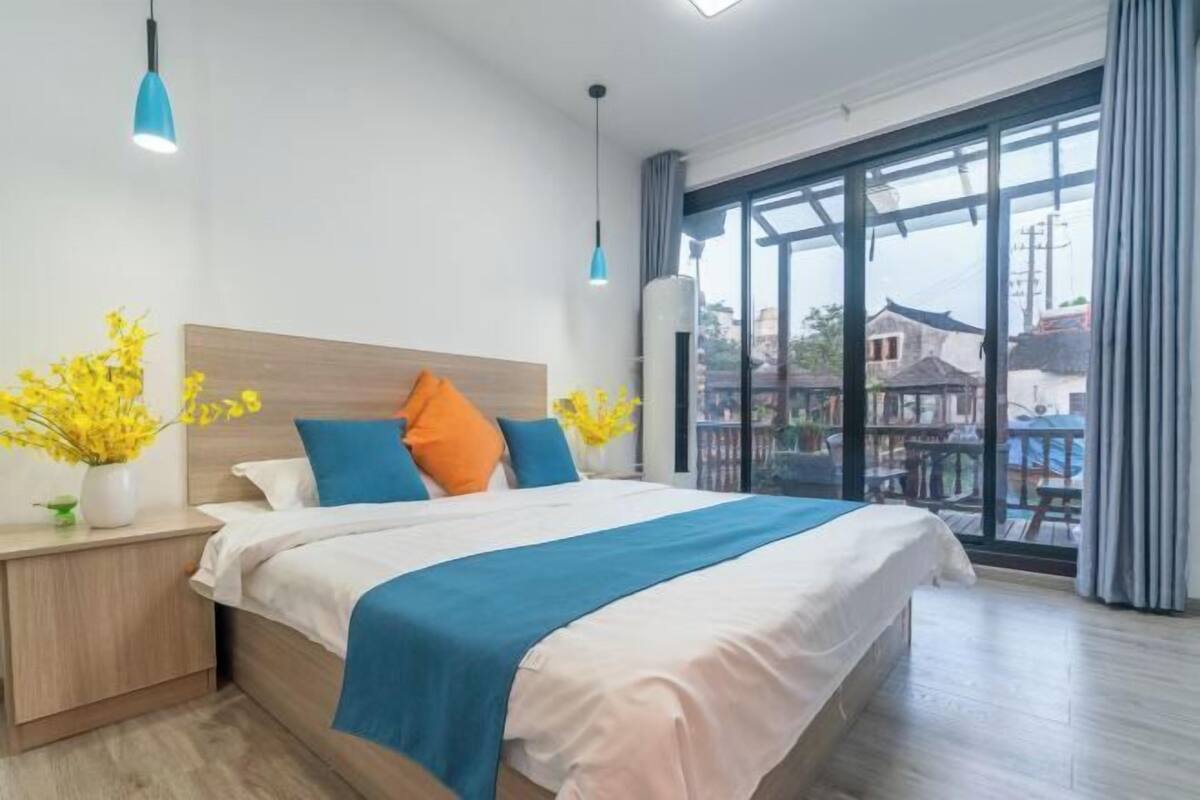 A modern hotel room with a large bed, a nightstand, a vase with yellow flowers, and a view of buildings through large windows.
