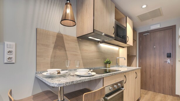A modern kitchen with granite countertops, stainless steel appliances, and wooden cabinetry. There are two bar stools and a hanging light fixture.