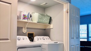 Una sala de bugaderia amb dues rentadores de càrrega frontal, una estant amb productes de neteja i una porta que condueix a una altra habitació.
