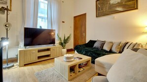 Un salon doté d’un téléviseur à écran plat, d’un canapé vert foncé, d’un fauteuil blanc, d’une table basse en bois et d’une grande fenêtre ornée de rideaux blancs.
