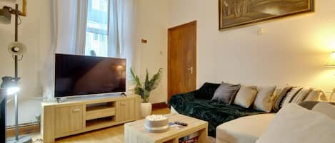 A living room with a flat-screen TV, a dark green sofa, a white armchair, a wooden coffee table, and a large window with white curtains.