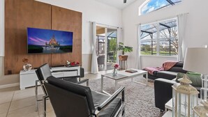 Ett rymligt vardagsrum med en platt-TV som är monterad på väggen, en takfläkt och stora fönster med utsikt över omgivningarna.