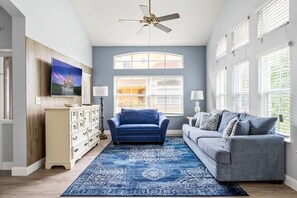 A living room with a blue sofa, a grey sofa, a patterned rug, a TV on the wall, and a ceiling fan. - Themed Bedrooms Near Disney 4BR (Kissimmee)