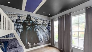 Una habitación con un mural de Star Wars, una cama con cubrecama gris y una ventana con cortinas grises.