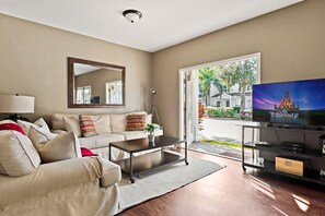 A living room with a beige sofa, a coffee table, a TV on a stand, a mirror, and a view of a house through a glass door. - Welcoming 4 BR Sleeps 10 Guests Wl2901 (Kissimmee)