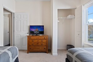 Une chambre avec une télévision posée sur une commode en bois, un lit recouvert d’une couette grise et blanche, et une fenêtre donnant sur l’extérieur.