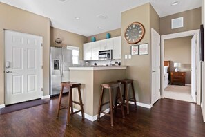 Una cocina moderna con armarios blancos, un refrigerador de acero inoxidable y un microondas. Hay tres taburetes de madera y un reloj de pared grande.