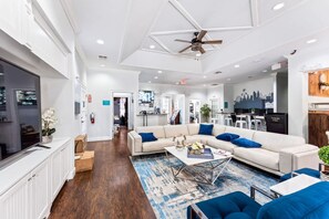Un vaste espace de vie comprenant un grand canapé d’angle, une table basse et un ventilateur de plafond. Au fond, on aperçoit une cuisine, et le mur est décoré d’une fresque représentant un paysage urbain.