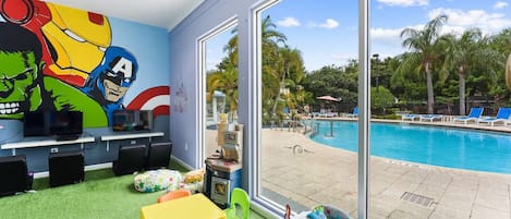 A children's play area with a pool view, featuring a mural of superheroes, play tables, and toys.
