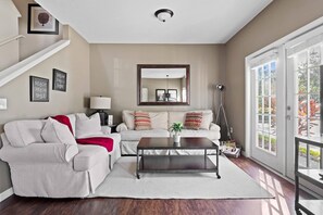 Un salon avec un canapé, une table basse et un grand miroir. On y trouve un escalier avec une rampe blanche et une porte donnant sur l’extérieur.