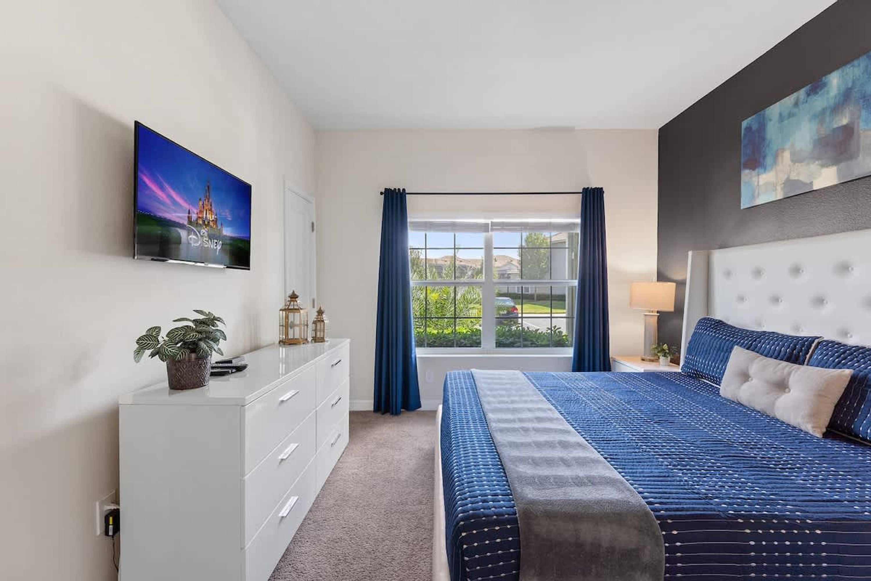 A modern bedroom with a large bed, a TV mounted on the wall, a dresser, and a window with curtains.