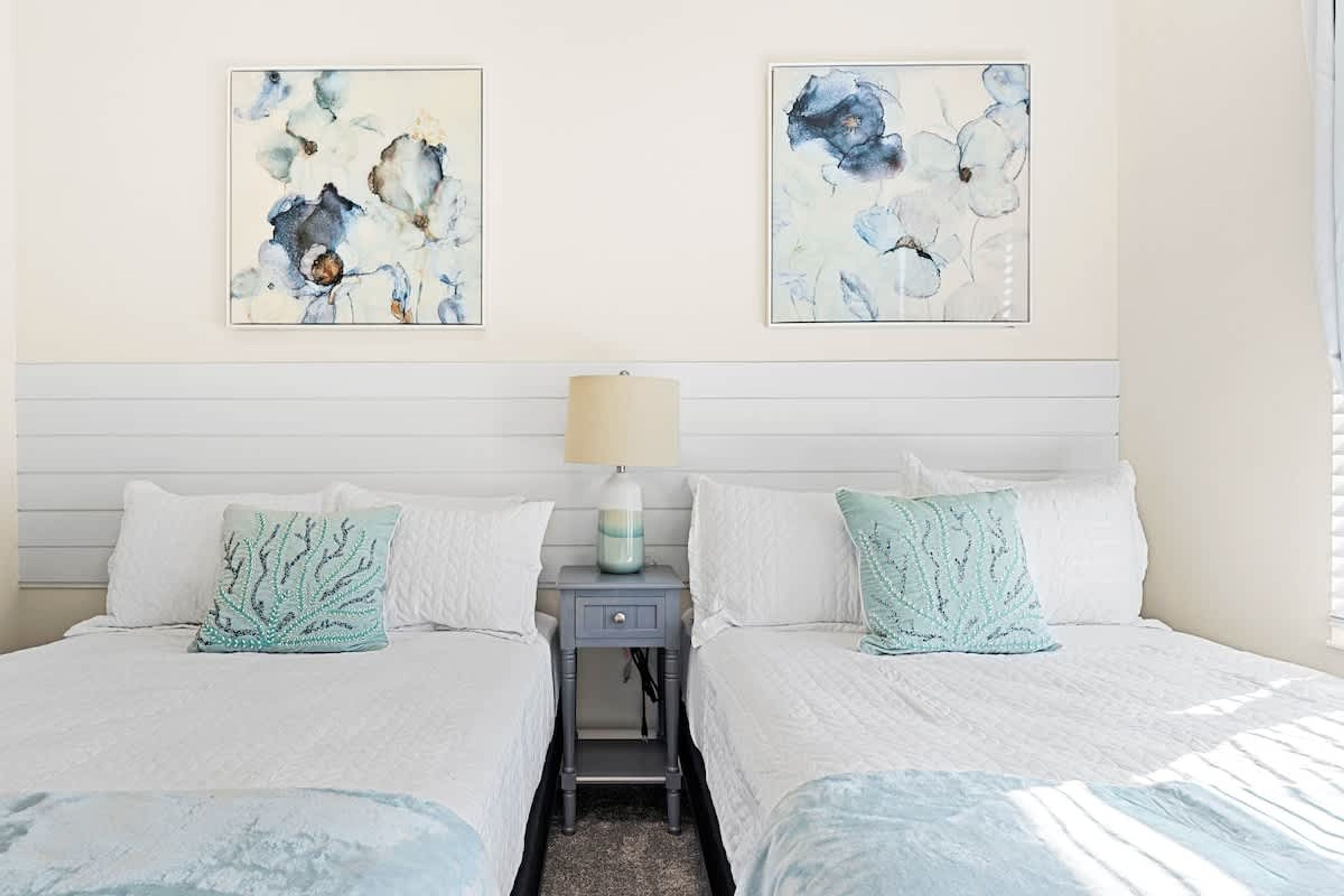 A bedroom with two beds, each with a white bedspread and a teal coral-patterned pillow. There is a nightstand with a lamp between the beds.