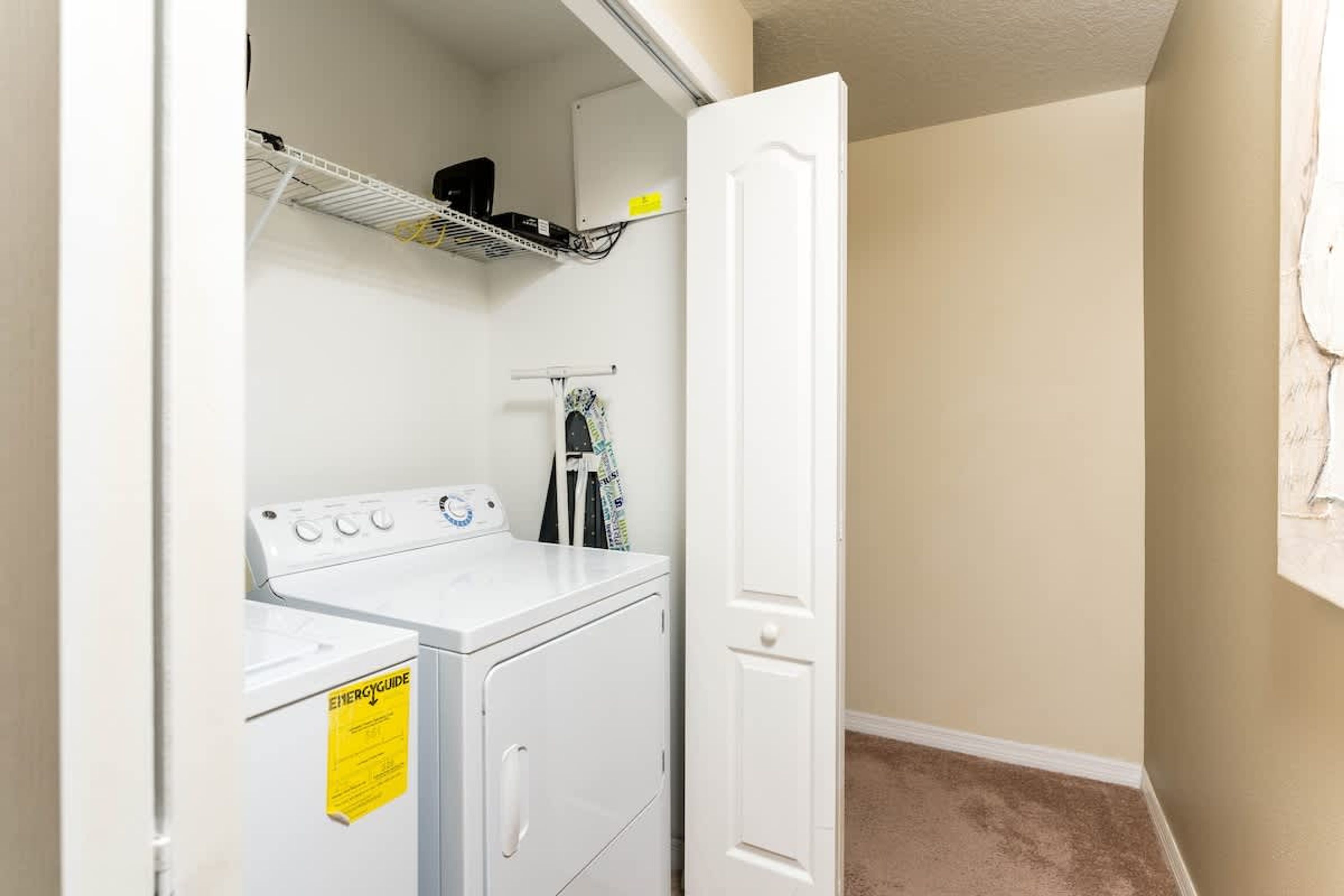A laundry room with a white washer and dryer, a shelf above the dryer, and a door to another room.