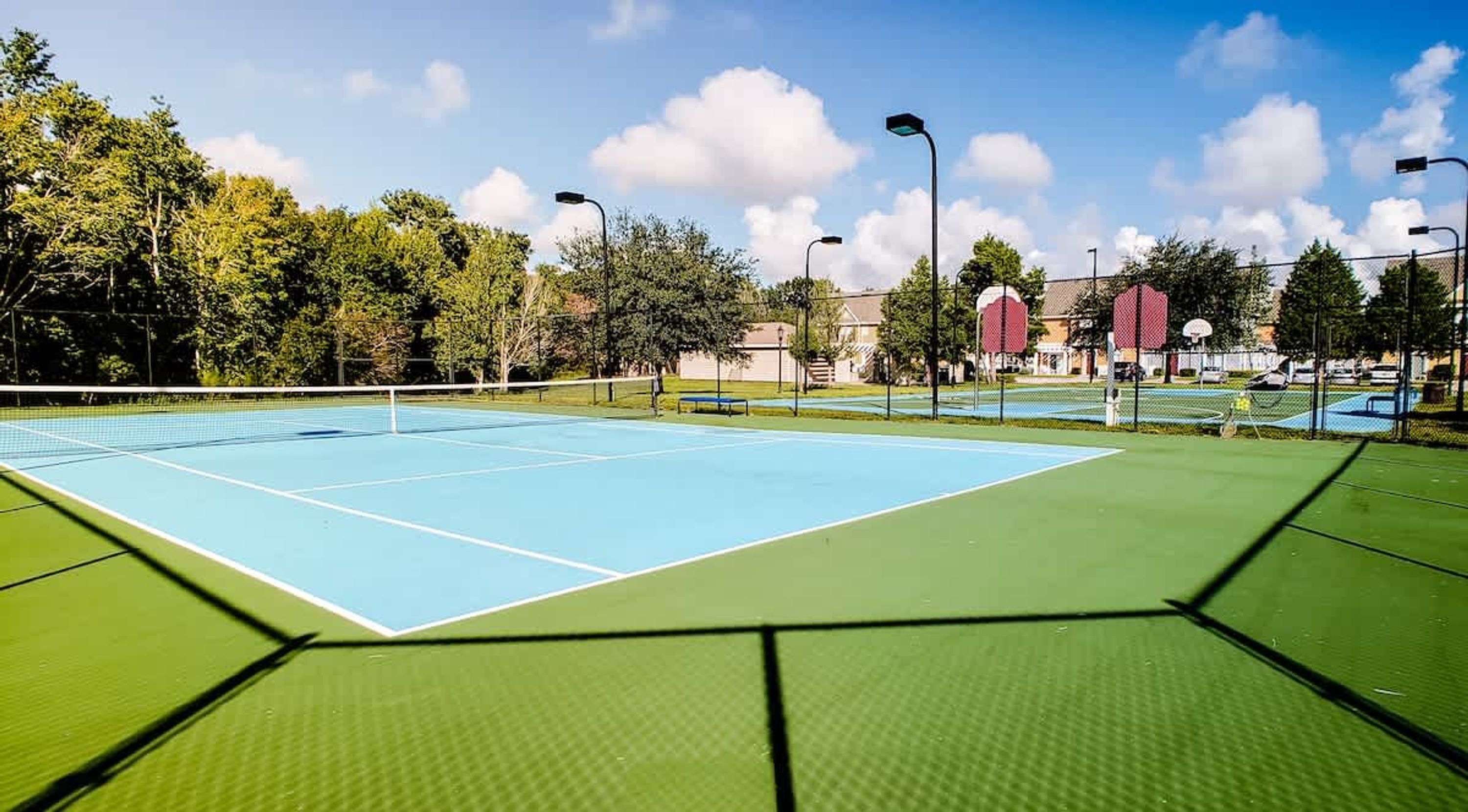 An outdoor tennis court with a green surface, surrounded by trees and a fence.