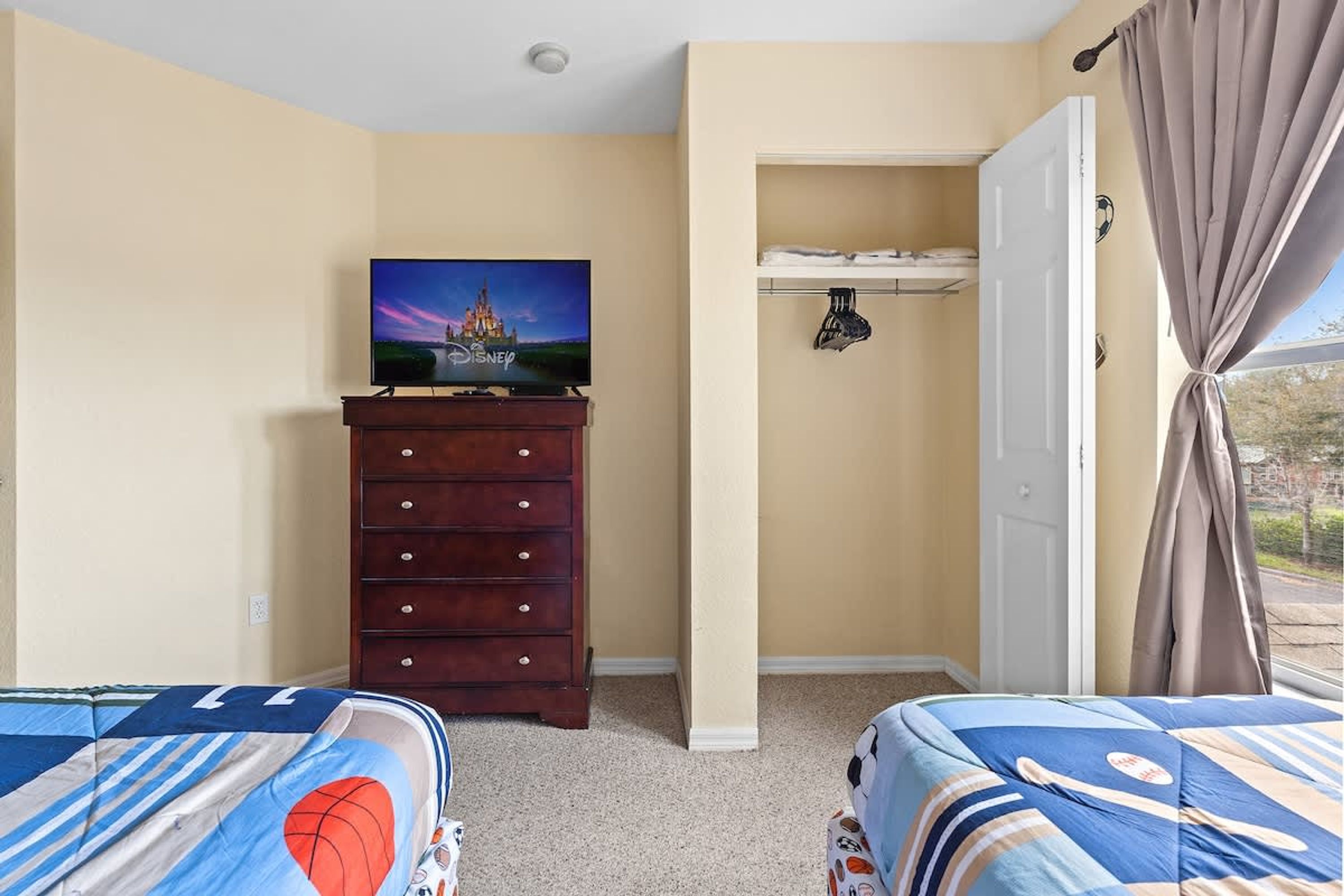 A bedroom with two beds, a dresser, a TV displaying a Disney castle, a closet, and a window with curtains.