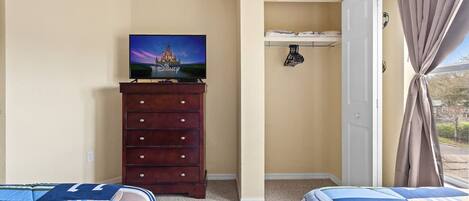 A bedroom with two beds, a dresser, a TV displaying a Disney castle, a closet, and a window with curtains.