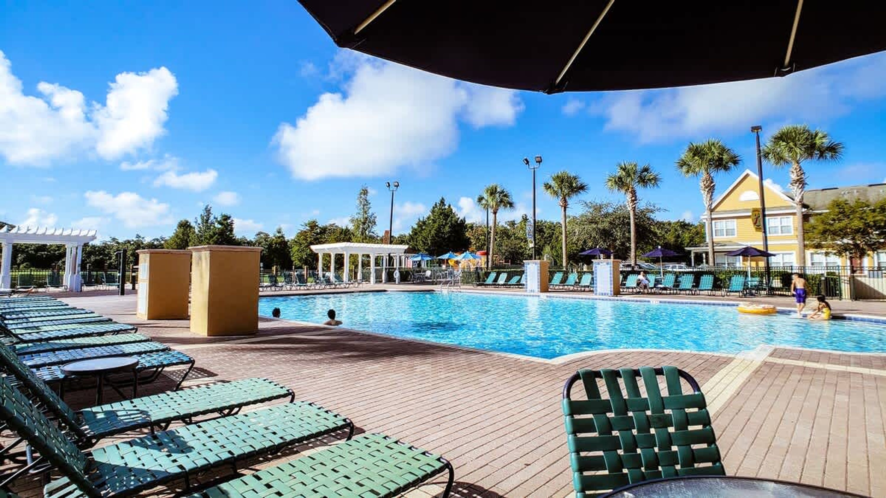 A swimming pool with lounge chairs and umbrellas, surrounded by palm trees and a clear sky.