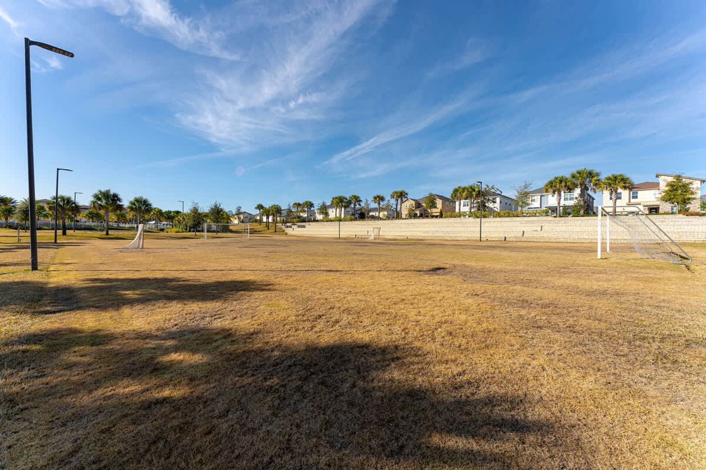 A wide, open field with a goalpost, surrounded by houses and palm trees under a clear sky.