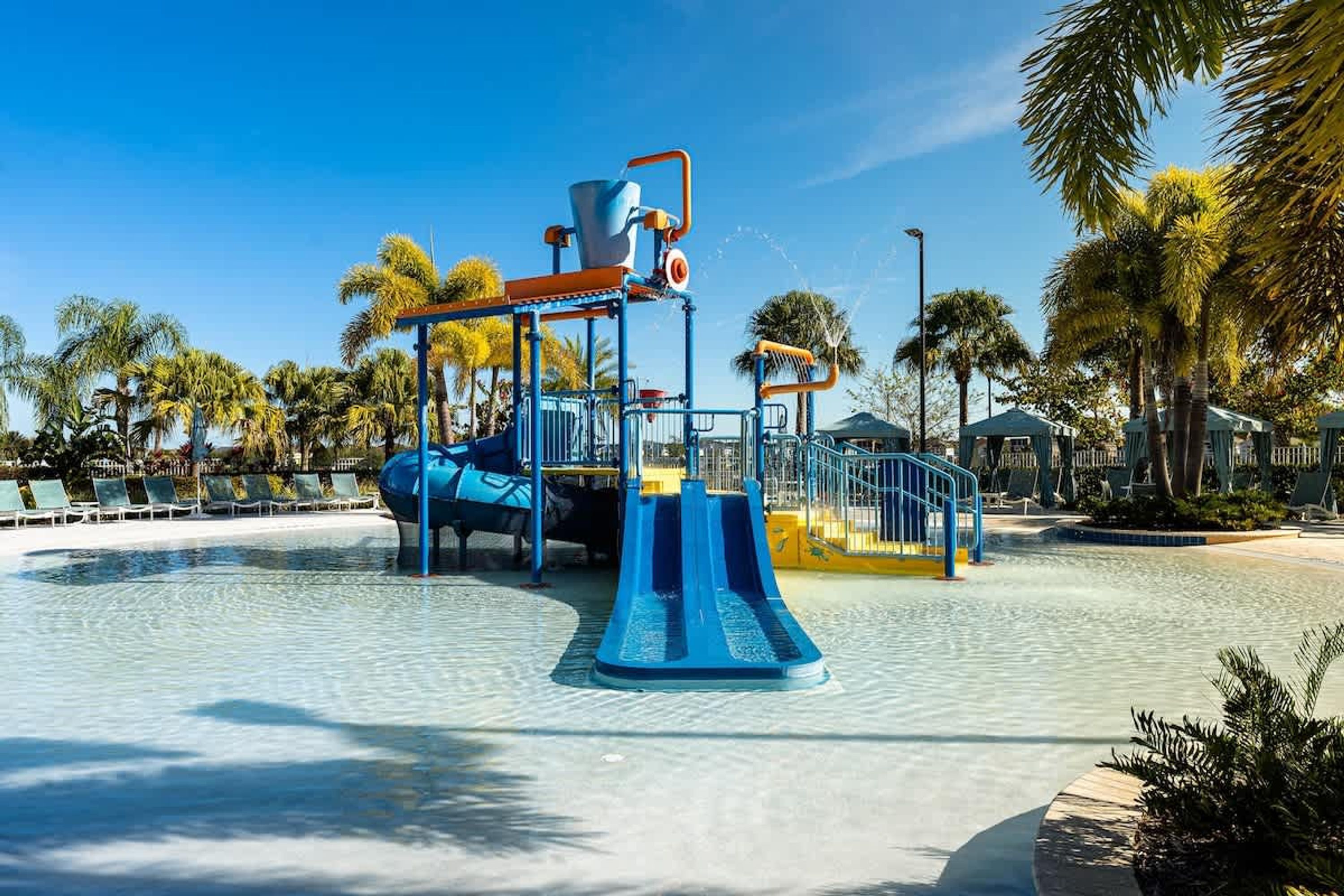 A water play area with slides and a water fountain, surrounded by palm trees and lounge chairs.