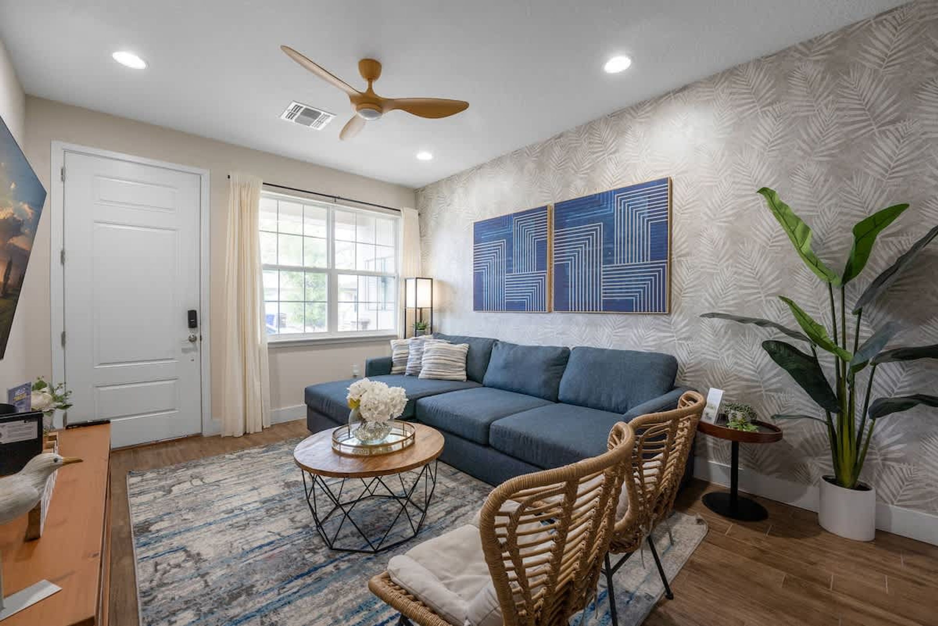 A living room with a sofa, a coffee table, a wicker chair, a ceiling fan, and a large potted plant.