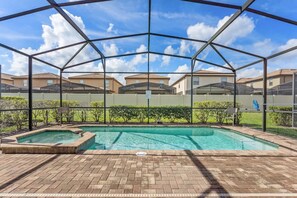 A covered outdoor swimming pool with a brick deck, surrounded by a metal fence and glass panels, with a view of residential buildings in the background. - Pet Friendly Amazing 5 BR Sleeps 10 Ww2116 (Kissimmee)