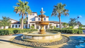 Ein mehrstufiger Brunnen, umgeben von einem kreisförmigen Steinrondell, mit Palmen und einem Gebäude im Hintergrund.