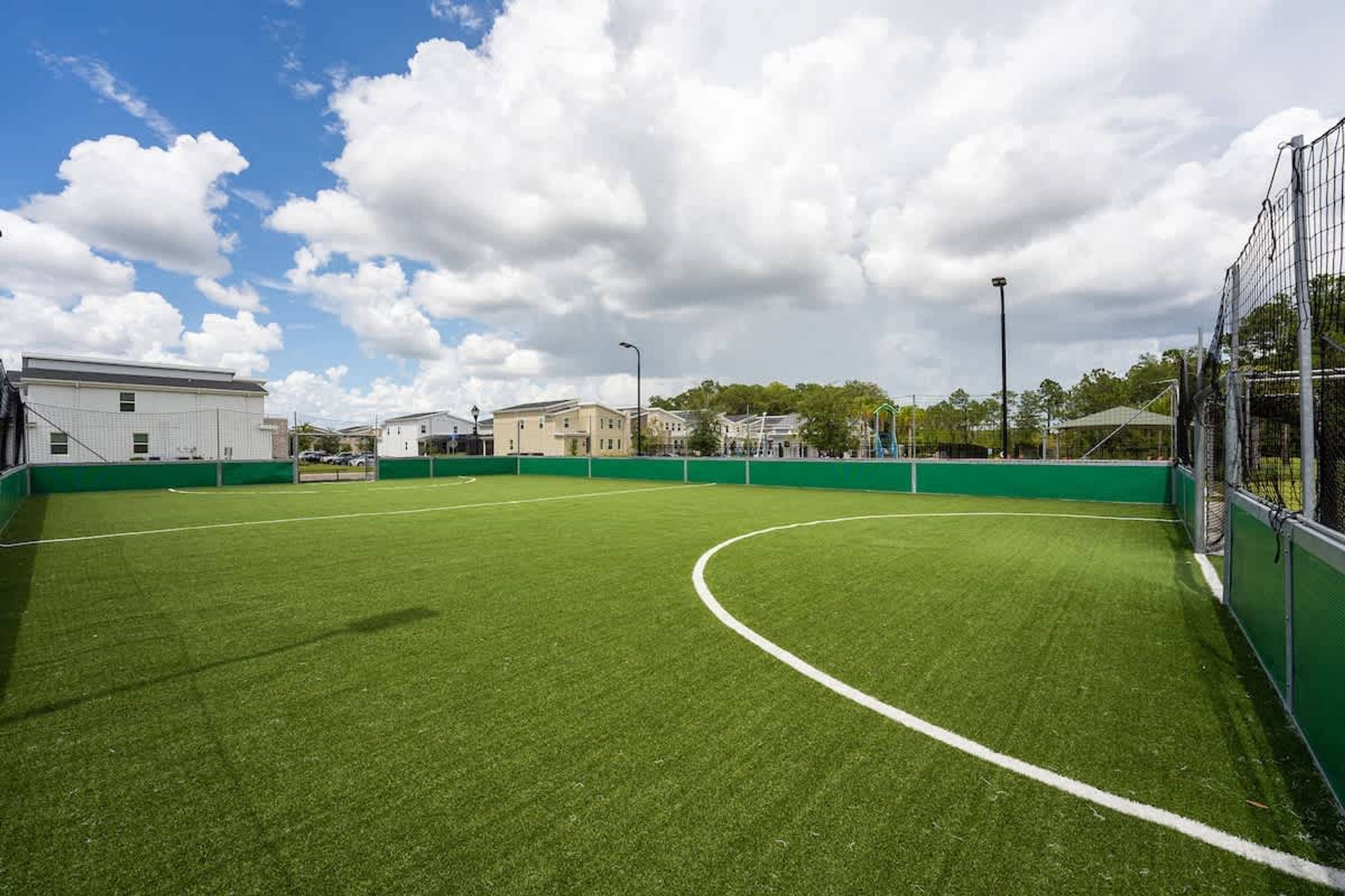 A soccer field with green artificial turf, surrounded by a green fence, and buildings in the background.