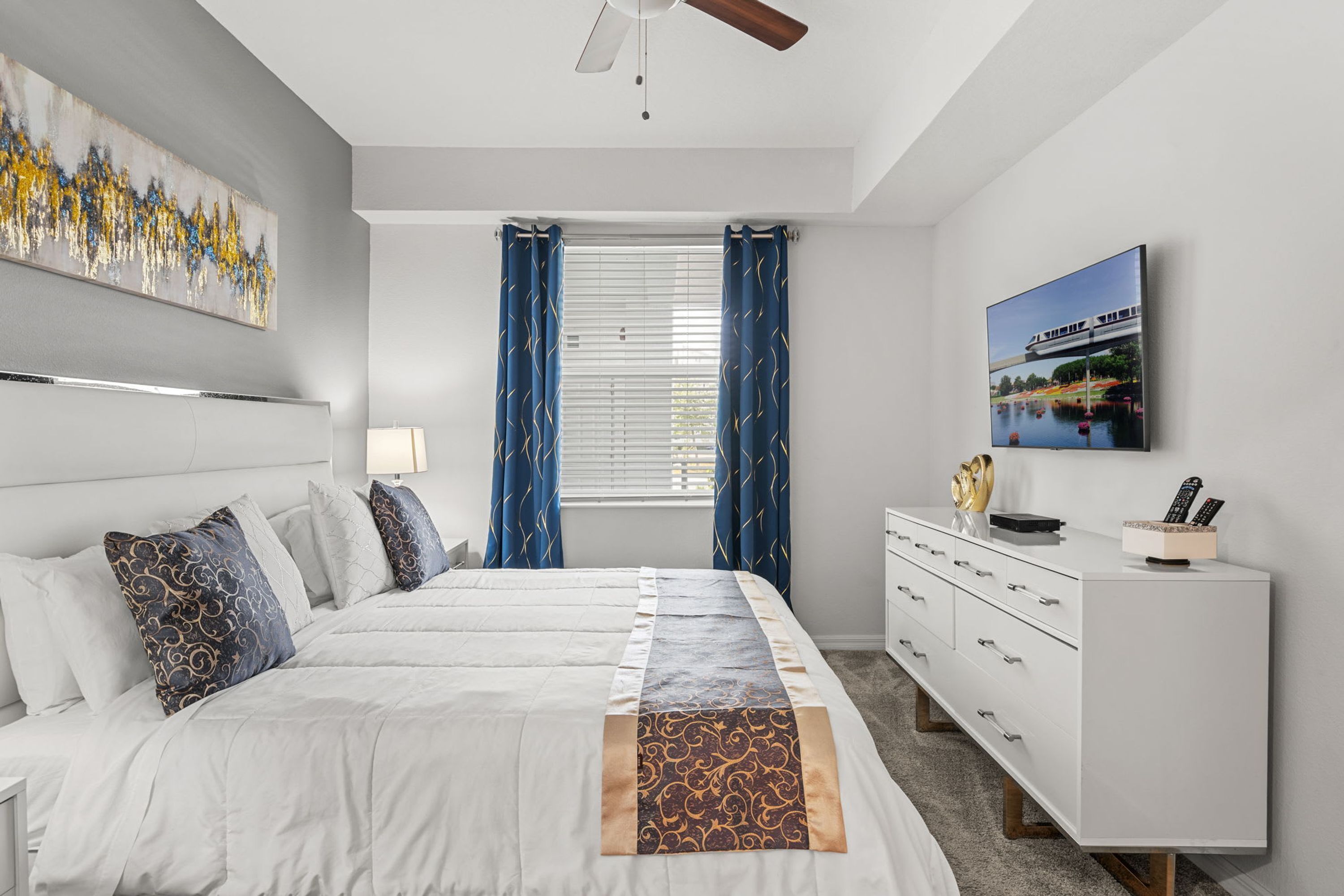 A bedroom with a large bed, a white dresser, a ceiling fan, a painting, and a window with curtains.