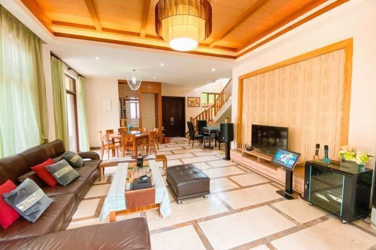 A spacious living room with a sofa, ottoman, and a flat-screen TV. There is a staircase leading to another level, and a dining area with chairs and a table.