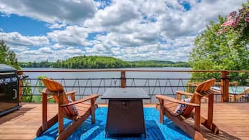 Relax on the deck with a serene view of the lake and surrounding greenery, featuring a comfortable seating area with a table and Adirondack chairs.