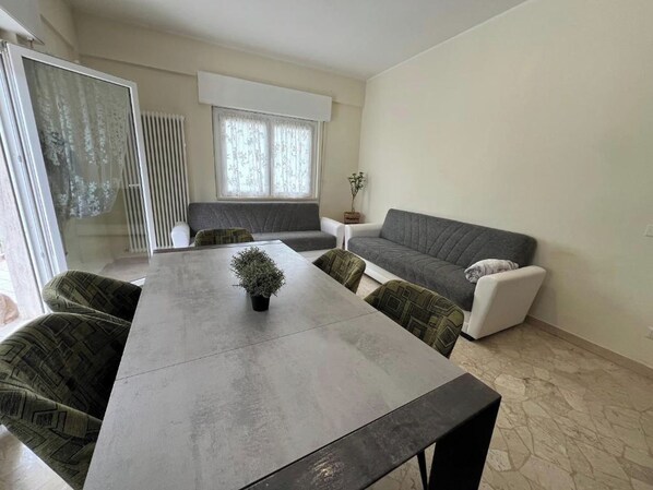 Ein modernes Wohnzimmer mit einem grauen Sofa, einem Esstisch mit vier Stühlen und einer Topfpflanze.