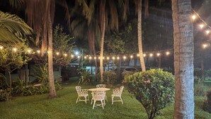 Un espace extérieur bien éclairé, avec une table et des chaises, entouré de palmiers et décoré de guirlandes lumineuses.