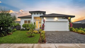A two-story house with a white exterior, a garage, and a well-maintained front yard with various plants and a small garden. - The Cozy Palm House (Homestead)