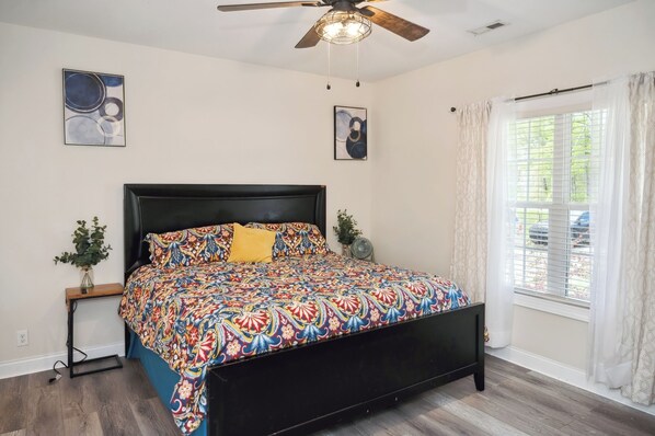 A bedroom with a black bed frame, colorful bedding, a ceiling fan, and a window with curtains. - Charming 3-bedroom house with AC, WiFi located in Supply NC near Holden Beach (supply)