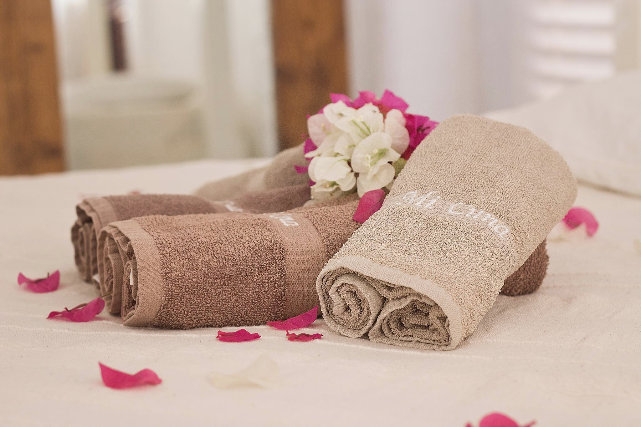 Toallas enrolladas con cuidado, adornadas con motivos florales y una etiqueta, sobre una cama con pétalos de rosa esparcidos.