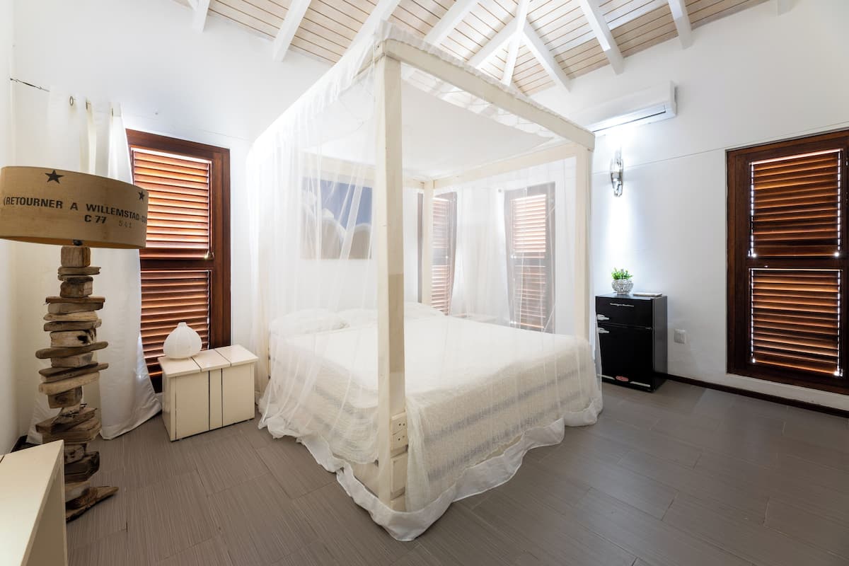 Una cama con dosel sobre cuatro columnas en una habitación con suelo de madera, una mesita de noche pequeña y un buró.