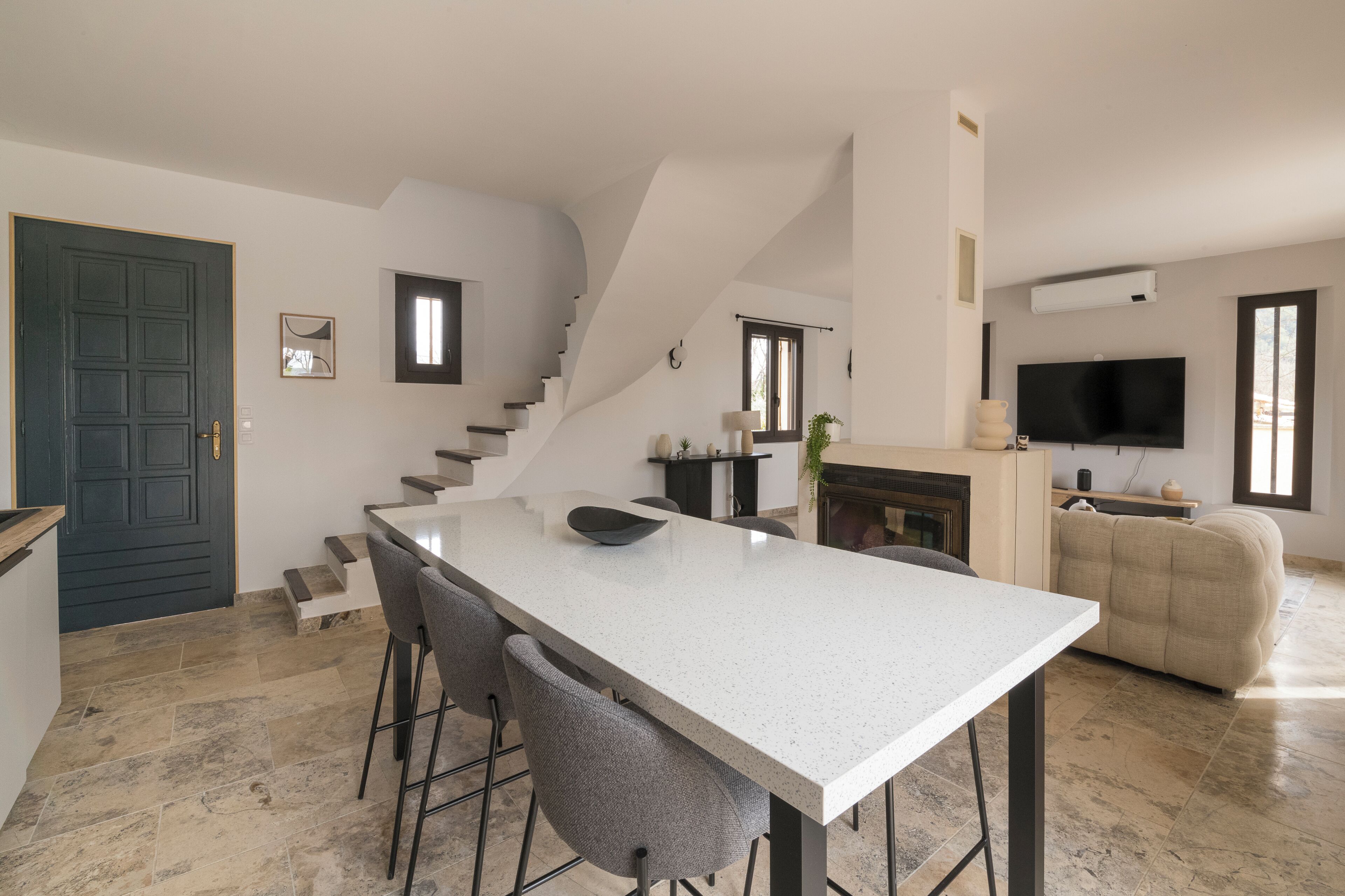 A modern kitchen with a central island, bar stools, a staircase, a flat-screen TV, and a fireplace.