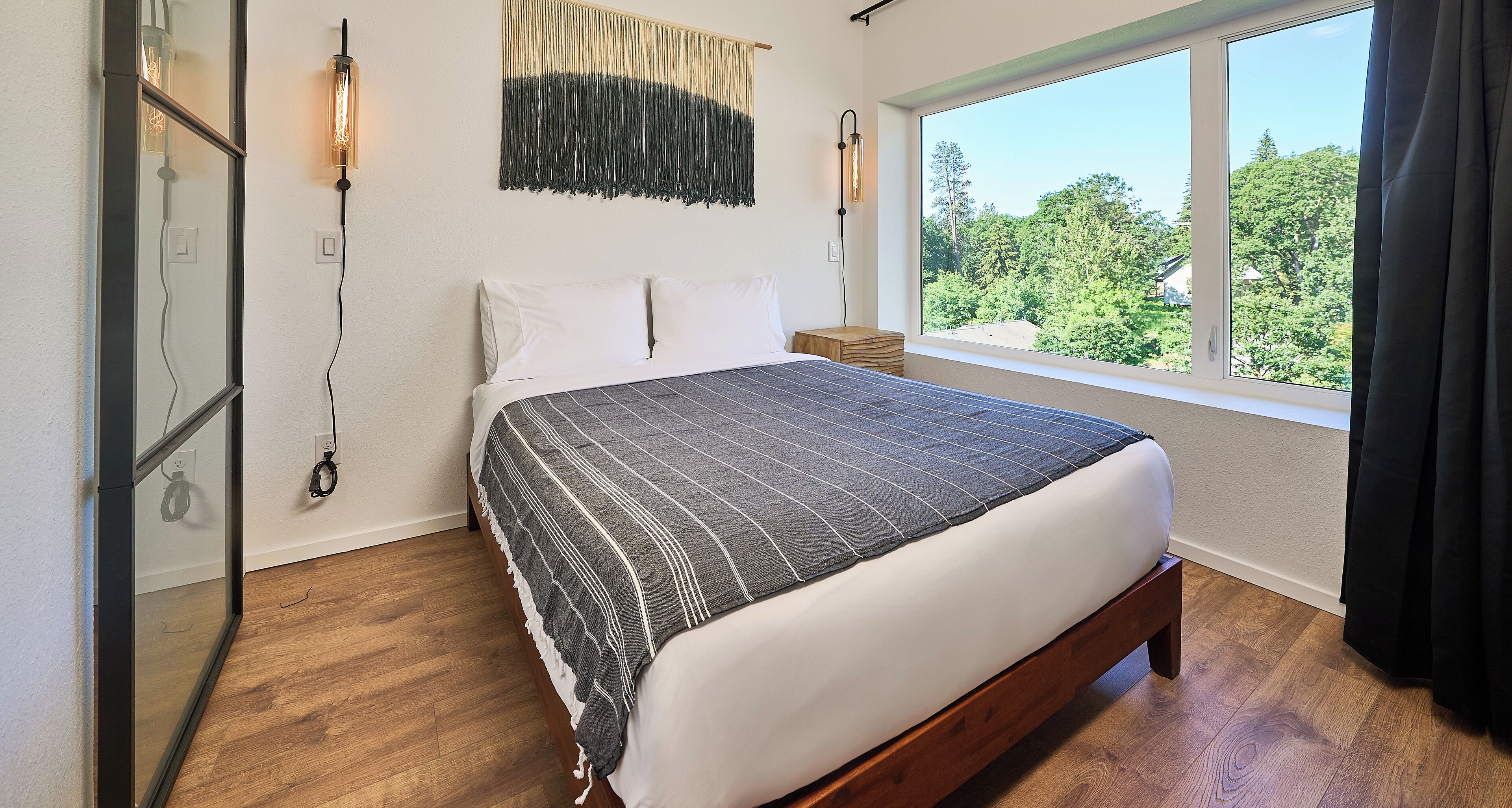A bedroom with a large bed, a view of trees through the window, and a wall-mounted light fixture.